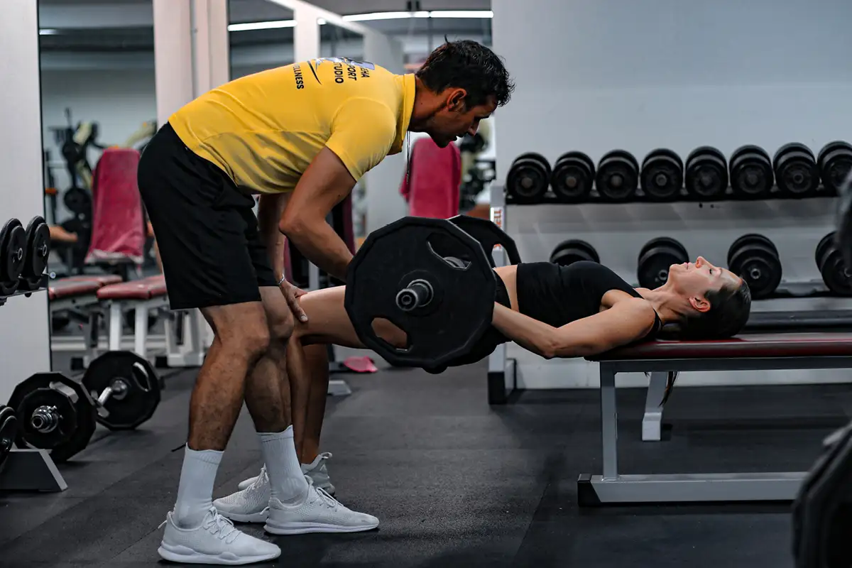 Trainer unterstützt Sportlerin beim Bankheben mit Langhantel im Fitnessstudio