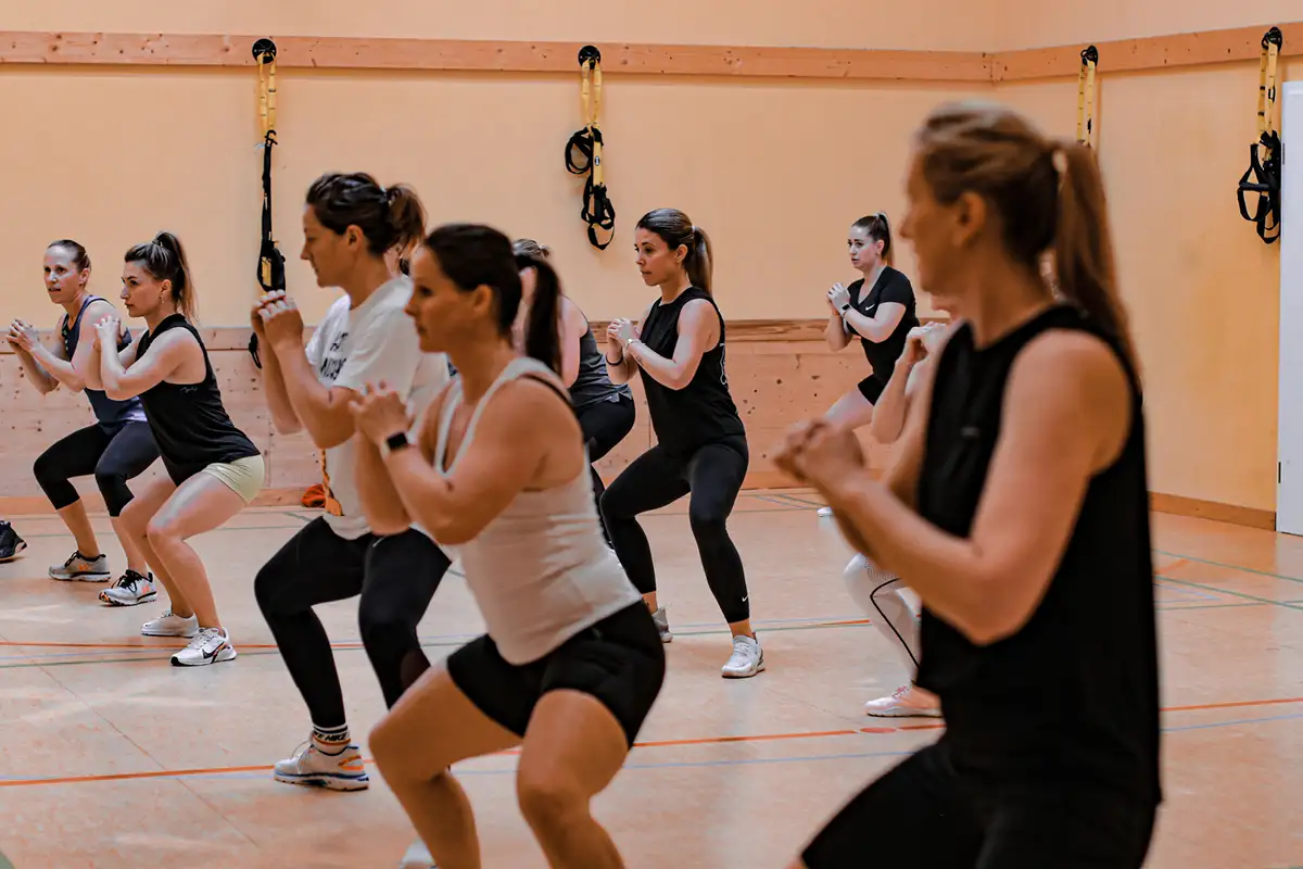 Gruppe beim Fitnesstraining im Kursraum — mehrere Personen in Hockstellung mit verschränkten Händen