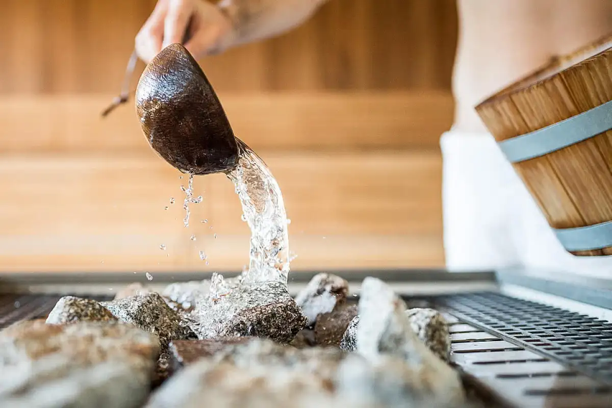 Nahaufnahme: Hand gießt mit einem Holzschöpfer Wasser auf heiße Saunasteine – Aufguss in einer Holzsauna.