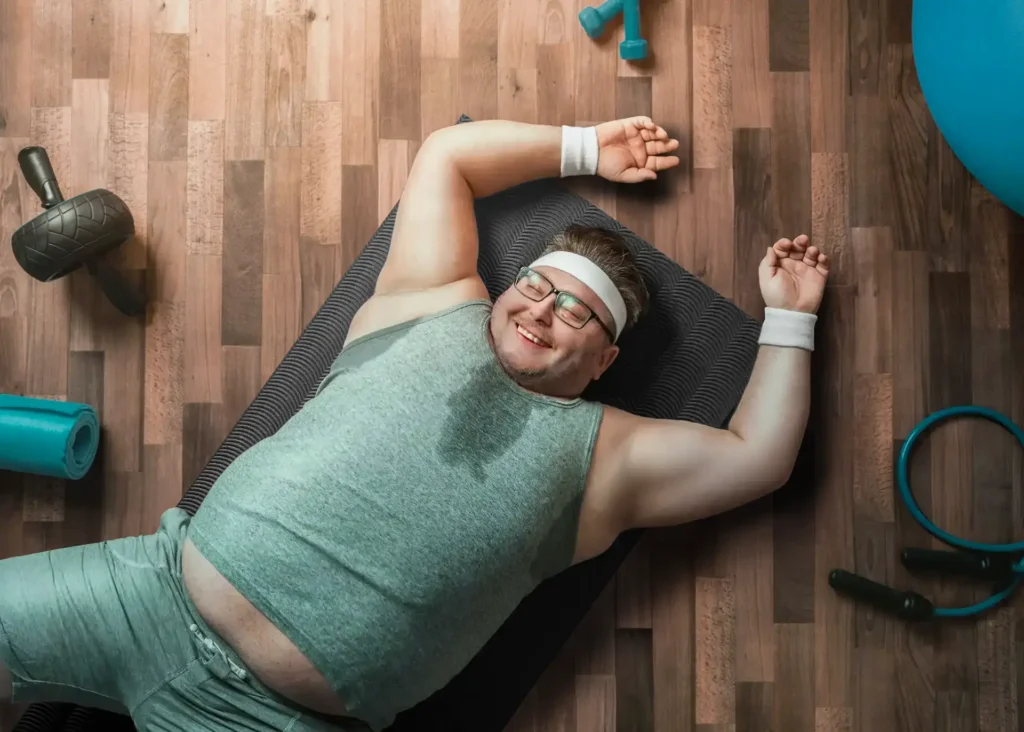 Lächelnde Person liegt nach dem Training erschöpft auf einer Matte; schweißnasses Shirt, Stirnband und Handgelenkbandagen; Trainingsgeräte liegen herum.