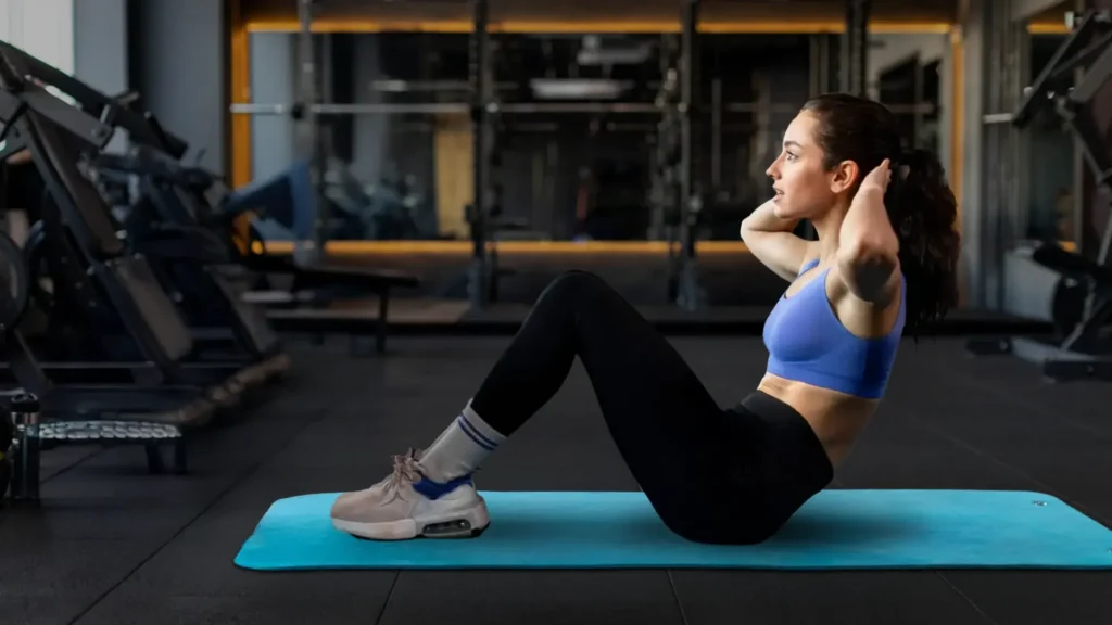 Frau macht Crunches auf einer blauen Matte im Fitnessstudio; Oberkörper angehoben, Hände an den Schläfen.