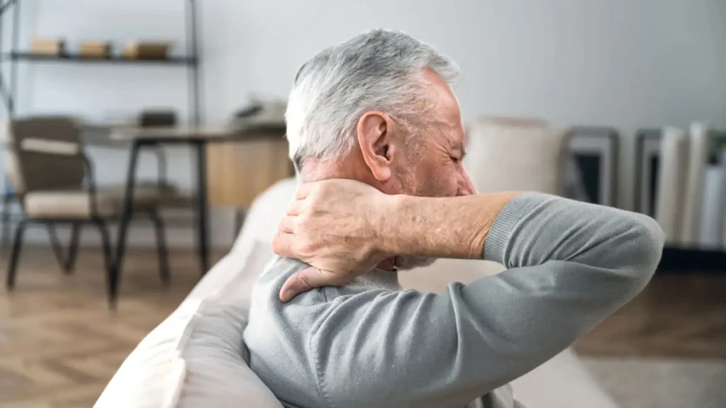 Älterer Mann sitzt auf dem Sofa und fasst sich schmerzend an den Nacken – Hinweis auf Nackenverspannungen.