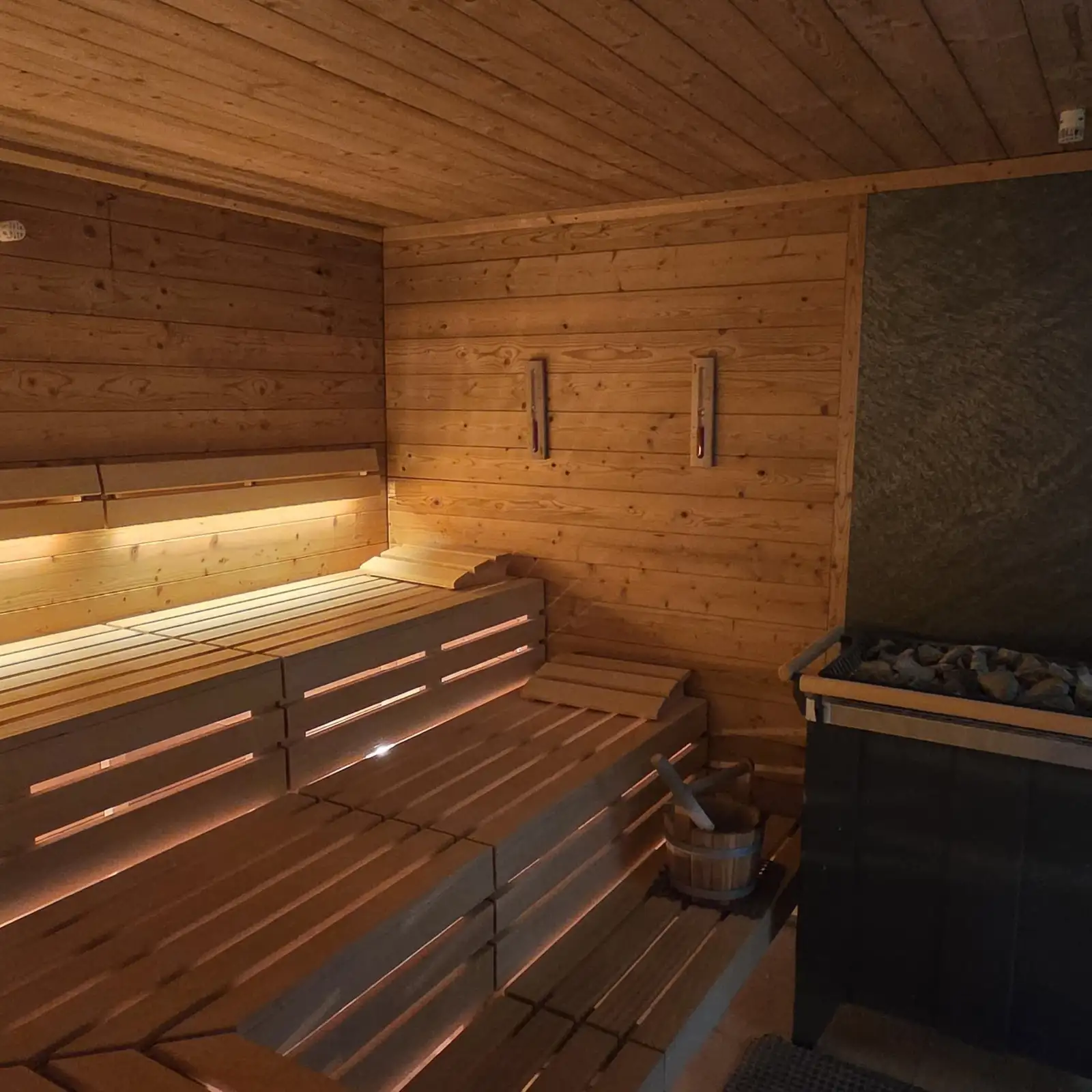 Sauna-Innenraum mit Holzbänken; Aufguss-Eimer und Schöpfer neben dem Ofen, warme Beleuchtung.
