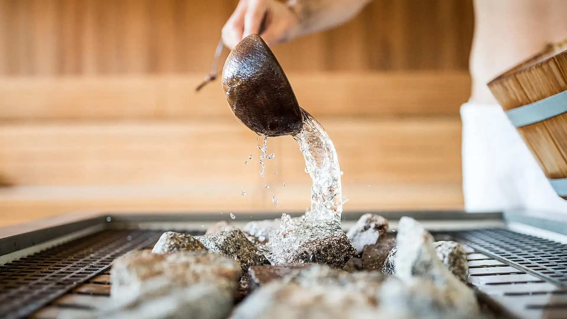 Nahaufnahme: Hand gießt mit einem Holzschöpfer Wasser auf heiße Saunasteine – Aufguss in einer Holzsauna.