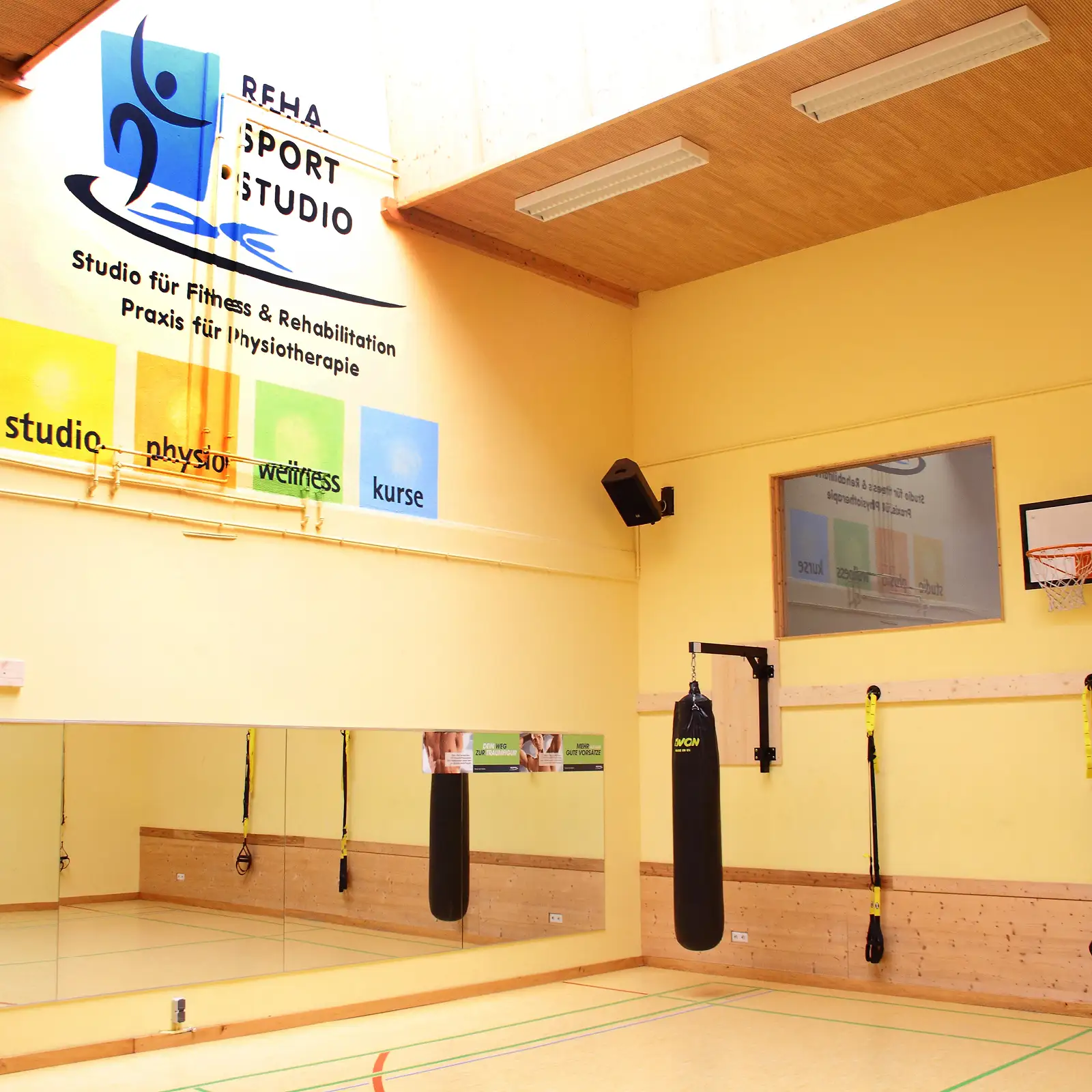 Heller Kursraum mit Spiegelwand, Boxsack und TRX-Schlingen; gelbe Wände mit Reha-Sportstudio-Logo.